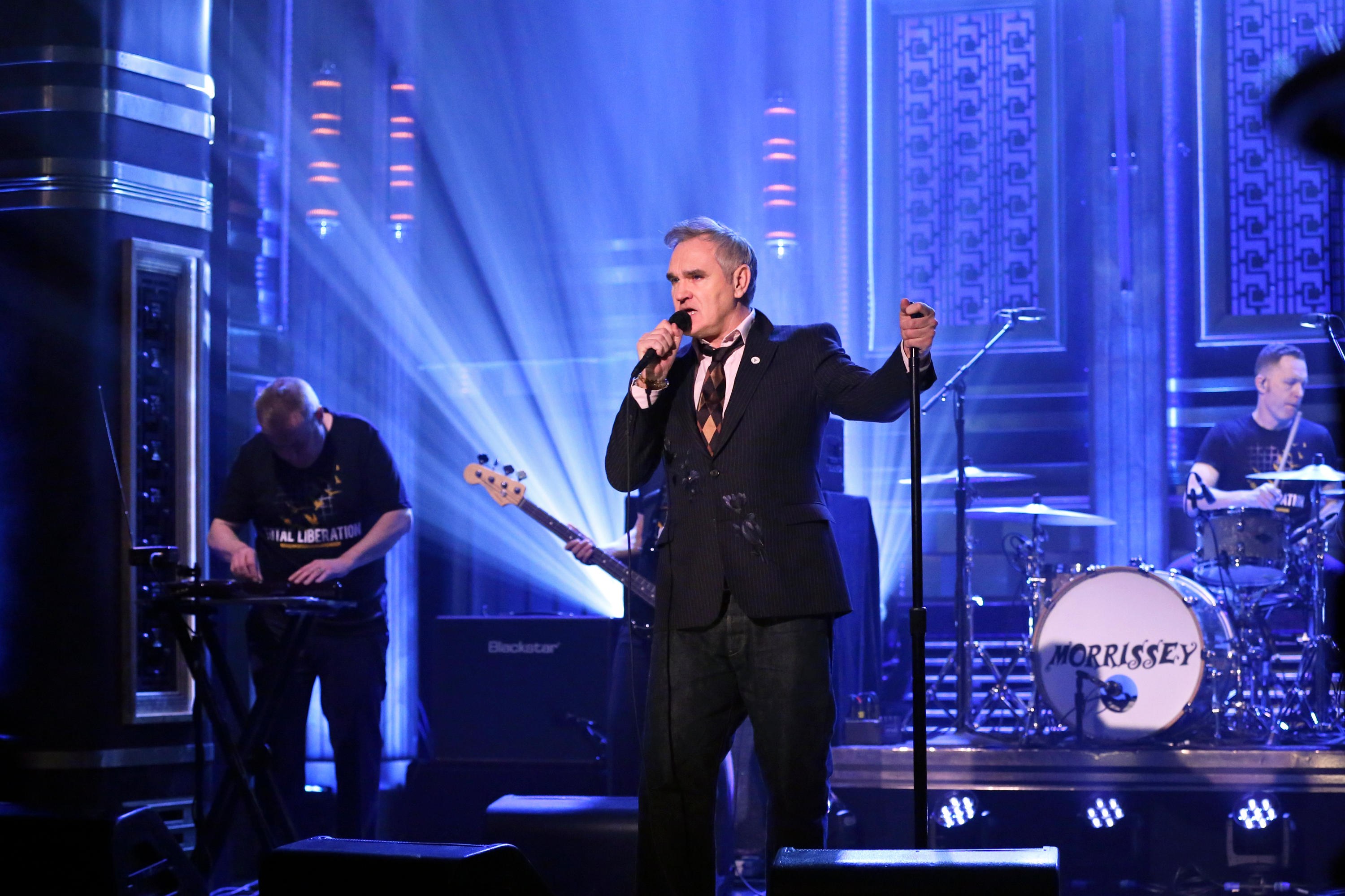 Morrissey performing on stage with his band under blue lights.
