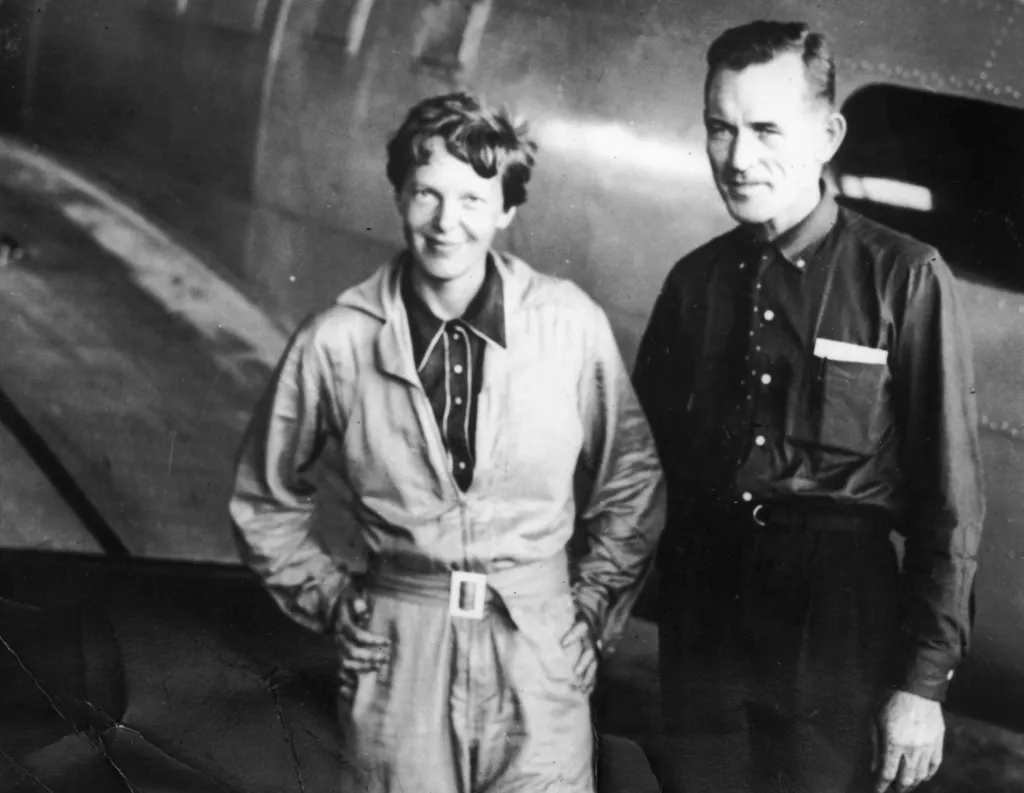 Amelia Earhart and Fred Noonan standing in a hangar, posing in front of an aircraft.