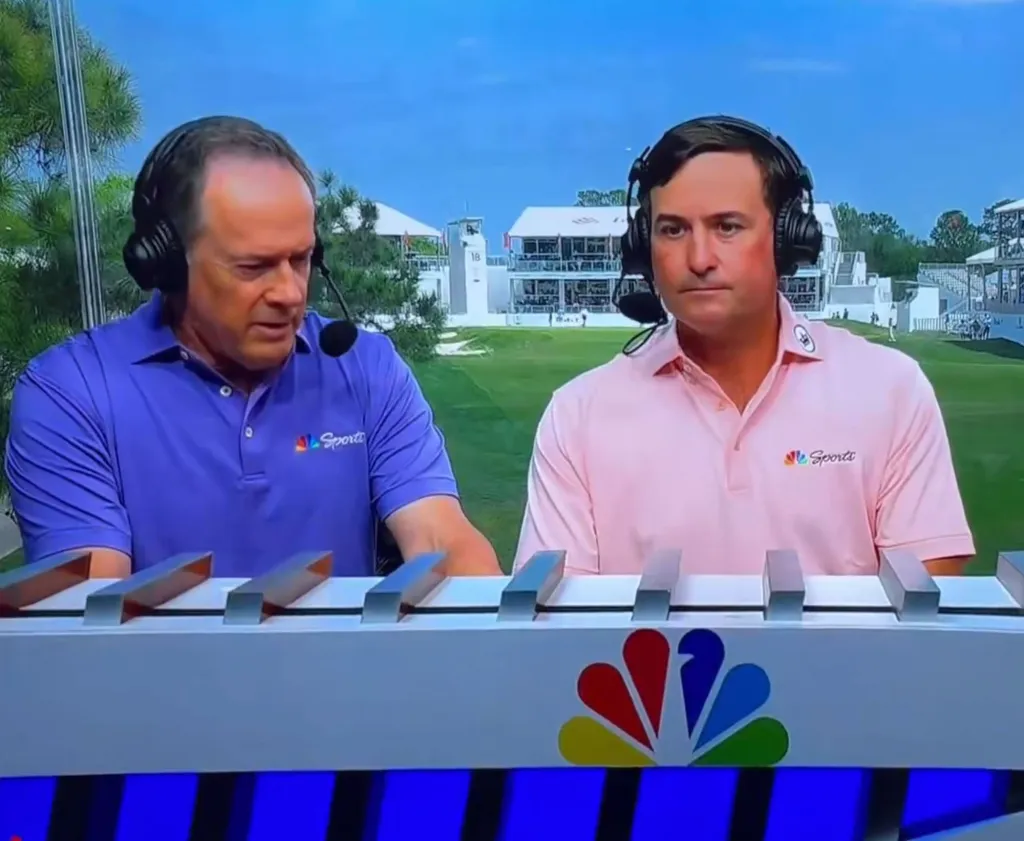 Two men in headsets and polo shirts, one purple and one pink, are sitting at a broadcast desk with the NBC Sports logo.
