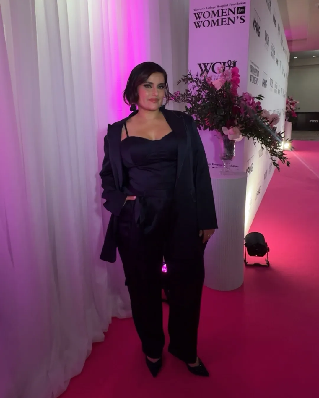 Nelly Furtado poses in a black plunging bodysuit and suit jacket on a pink carpet.