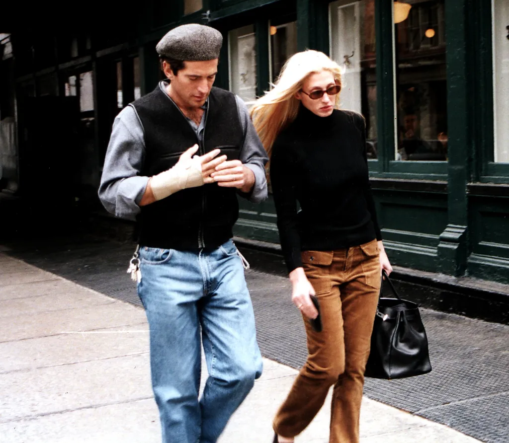 John F. Kennedy Jr. in a flat cap and Carolyn Bessette-Kennedy in sunglasses walking on a city street.