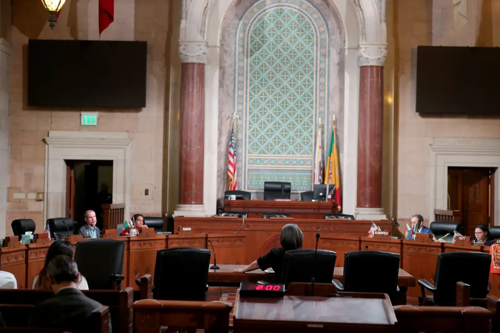 A person in black behind a wooden desk facing the front of the room where other people are seated at desks, during a Homelessness & Housing Committee meeting.