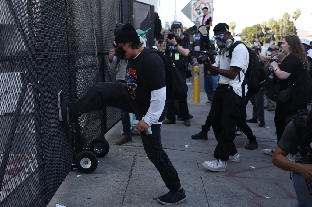 A person in a black beanie, t-shirt, and pants, with a black mask on their face, bracing a metal fence with their leg.