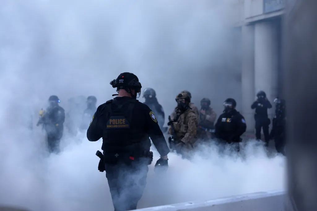 Law enforcement officers in riot gear stand in a cloud of white smoke.