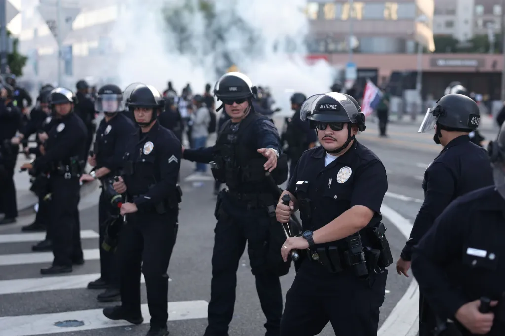 Several police officers in riot gear stand in a street with smoke in the background.