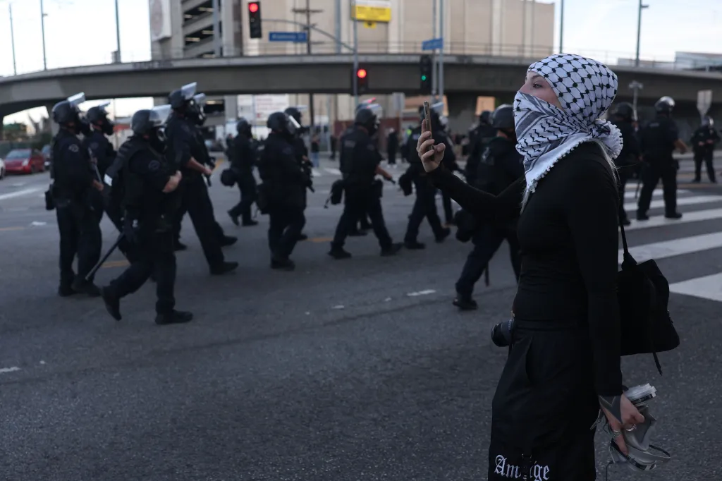 A person wearing a keffiyeh and holding a phone up to record a line of police officers marching in the street.