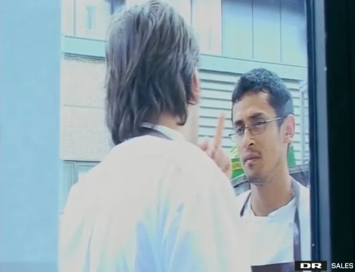 A person in a white chef's coat with their back to the viewer talks to another person in a white chef's coat and glasses who is wagging their finger.