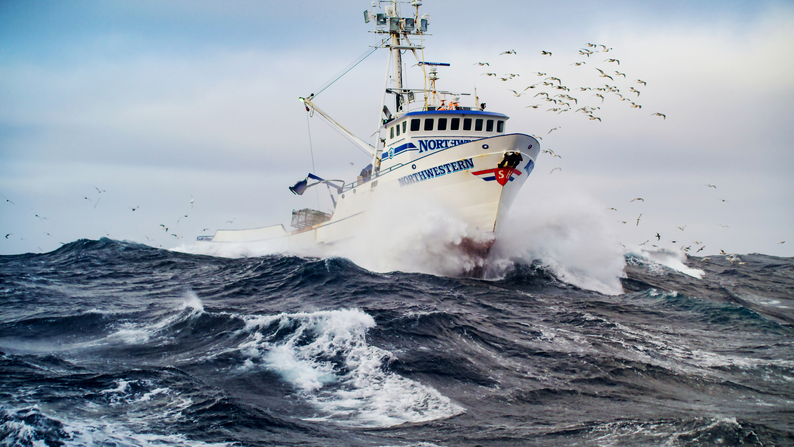 The fishing vessel Northwestern at sea, breaking through a wave as a flock of birds flies around the wheelhouse.