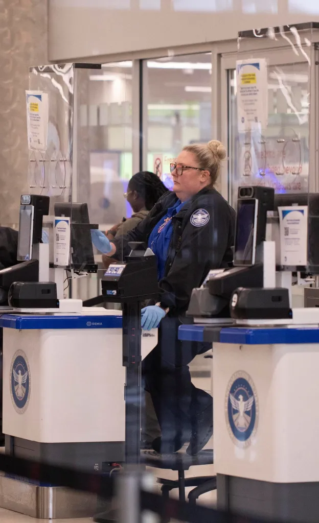 A TSA officer in glasses and a dark uniform sitting behind a security checkpoint.