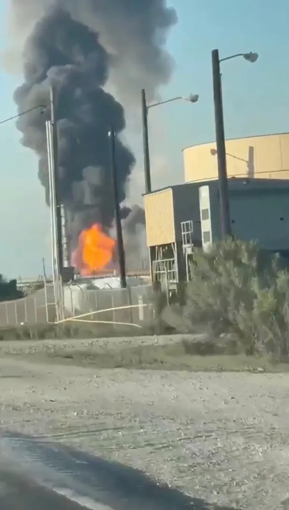 An oil refinery explosion with a large plume of black smoke and orange flames.