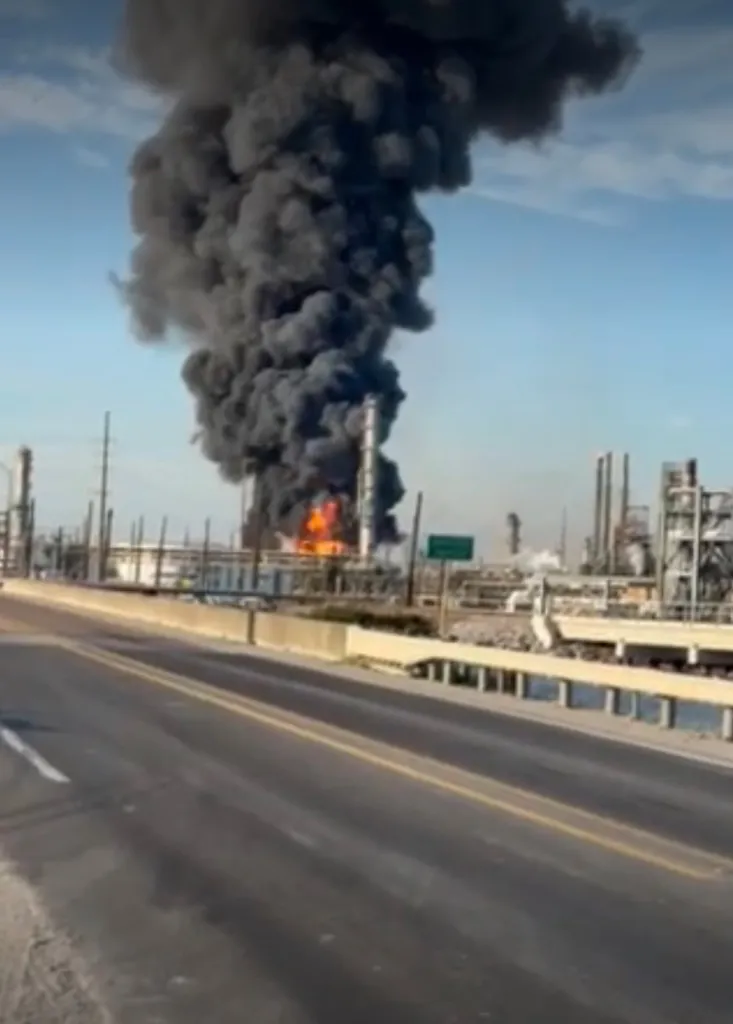 An oil refinery fire with a large plume of black smoke rising into the sky.