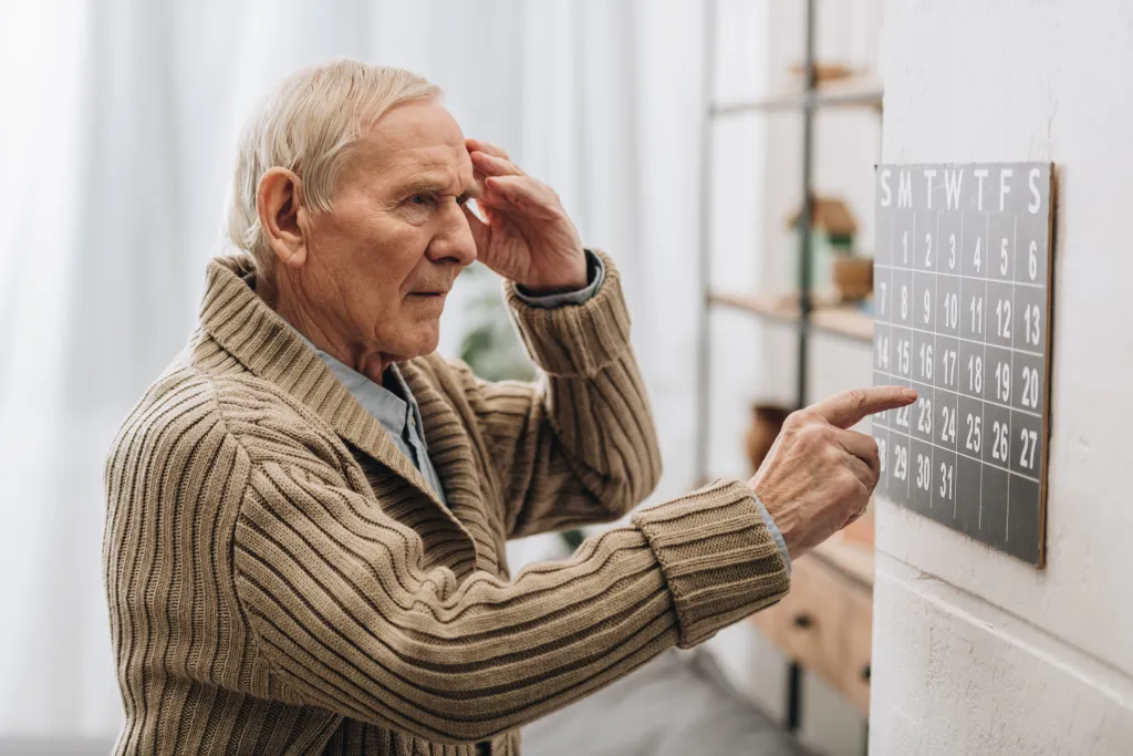 An elderly man touches his head in frustration while pointing to a wall calendar, suggesting memory loss.