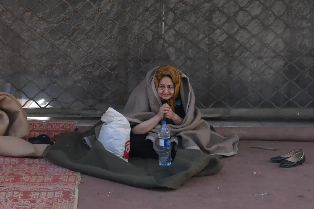 An old woman wrapped in a blanket sits on the ground, having been rescued from her damaged home in Tehran.