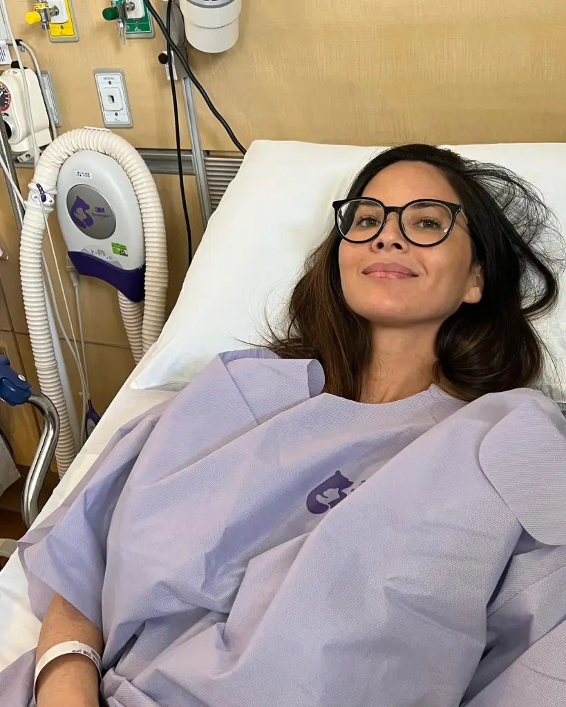 Olivia Munn lies in a hospital bed wearing a gown and glasses.