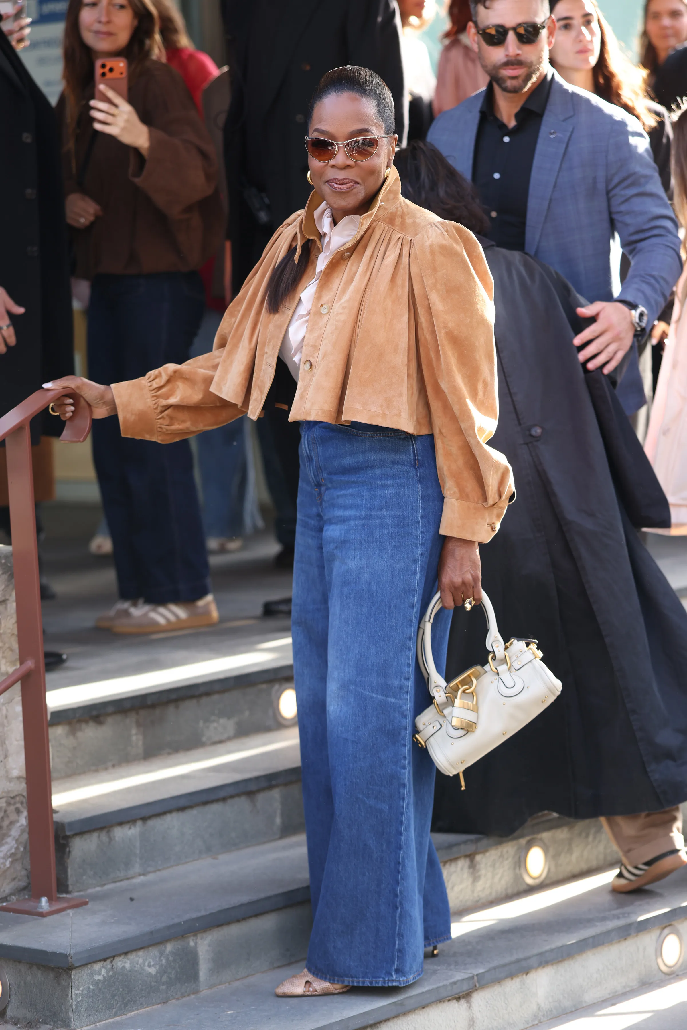 Oprah Winfrey attends Chloe during Paris Fashion Week, wearing a tan suede jacket, wide-leg jeans, and carrying a white handbag.
