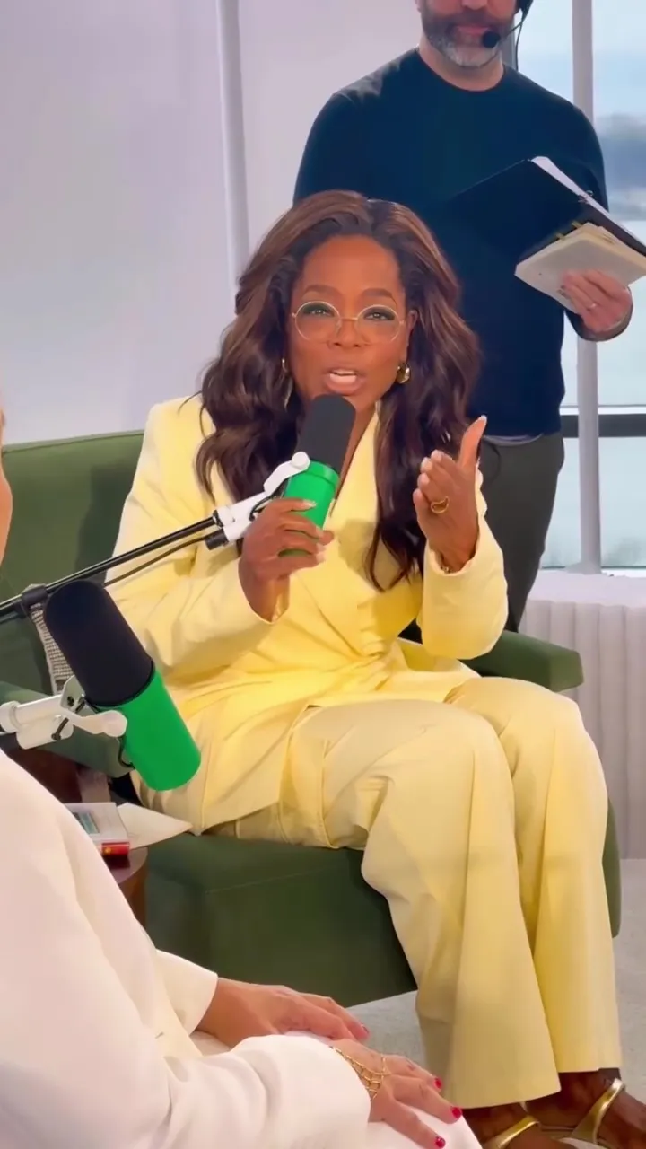 Oprah Winfrey in a yellow pantsuit, holding a green microphone and speaking during an interview.