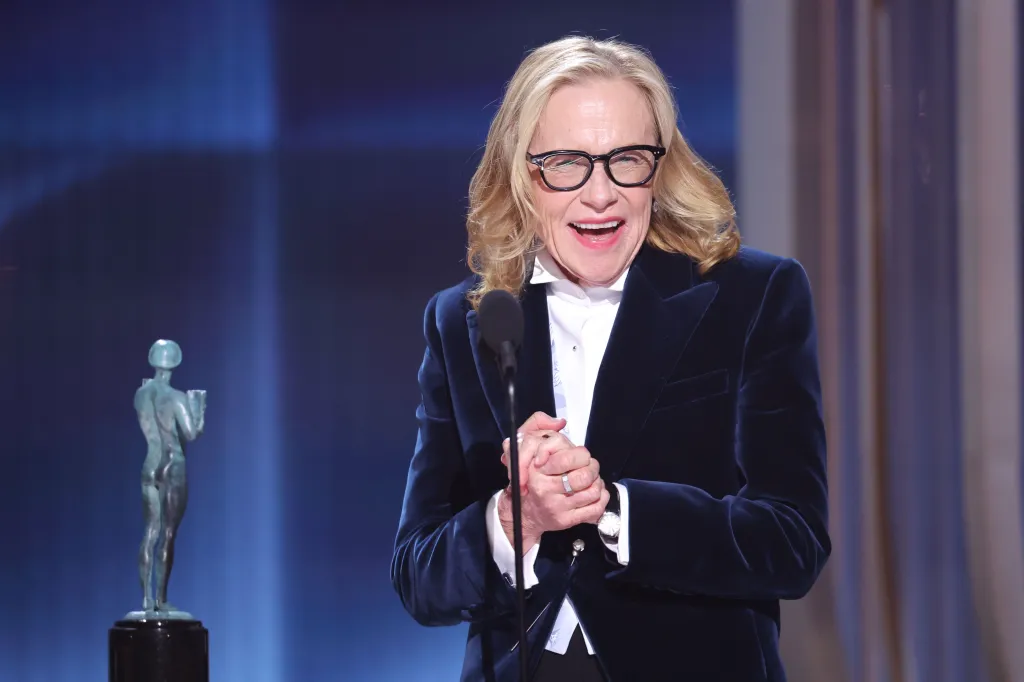 Amy Madigan smiling and wearing a dark blue velvet jacket and glasses, speaking at a microphone at the Actor Awards.