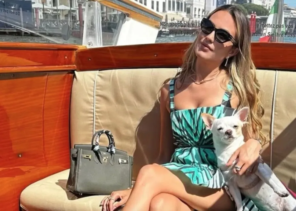 Pamela Genini, wearing sunglasses and a patterned dress, sits on a boat holding her white chihuahua, with a grey handbag next to her.
