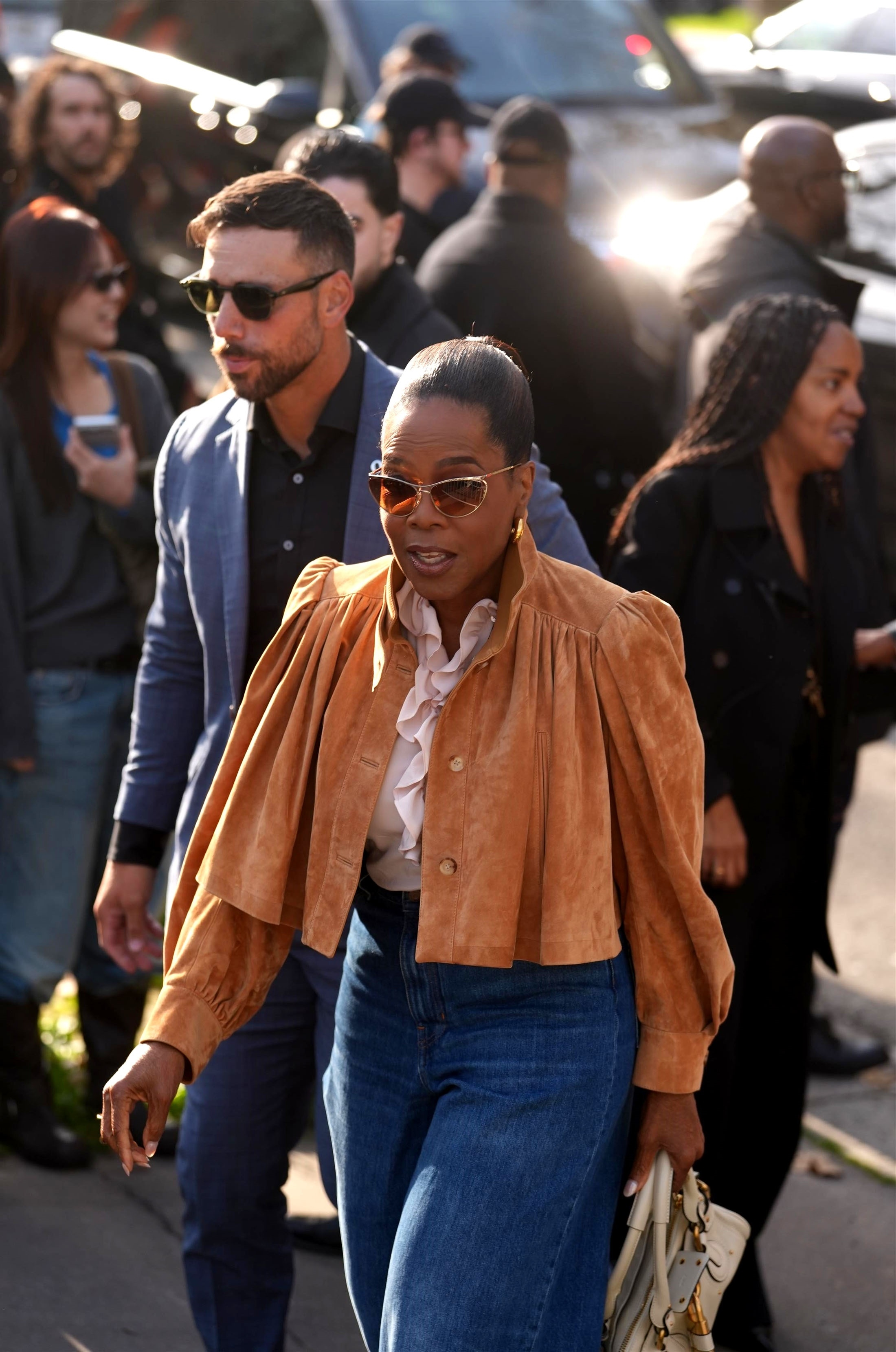 Oprah Winfrey arriving at the Chloe Fashion Show, wearing a brown suede jacket, blue jeans, and sunglasses.