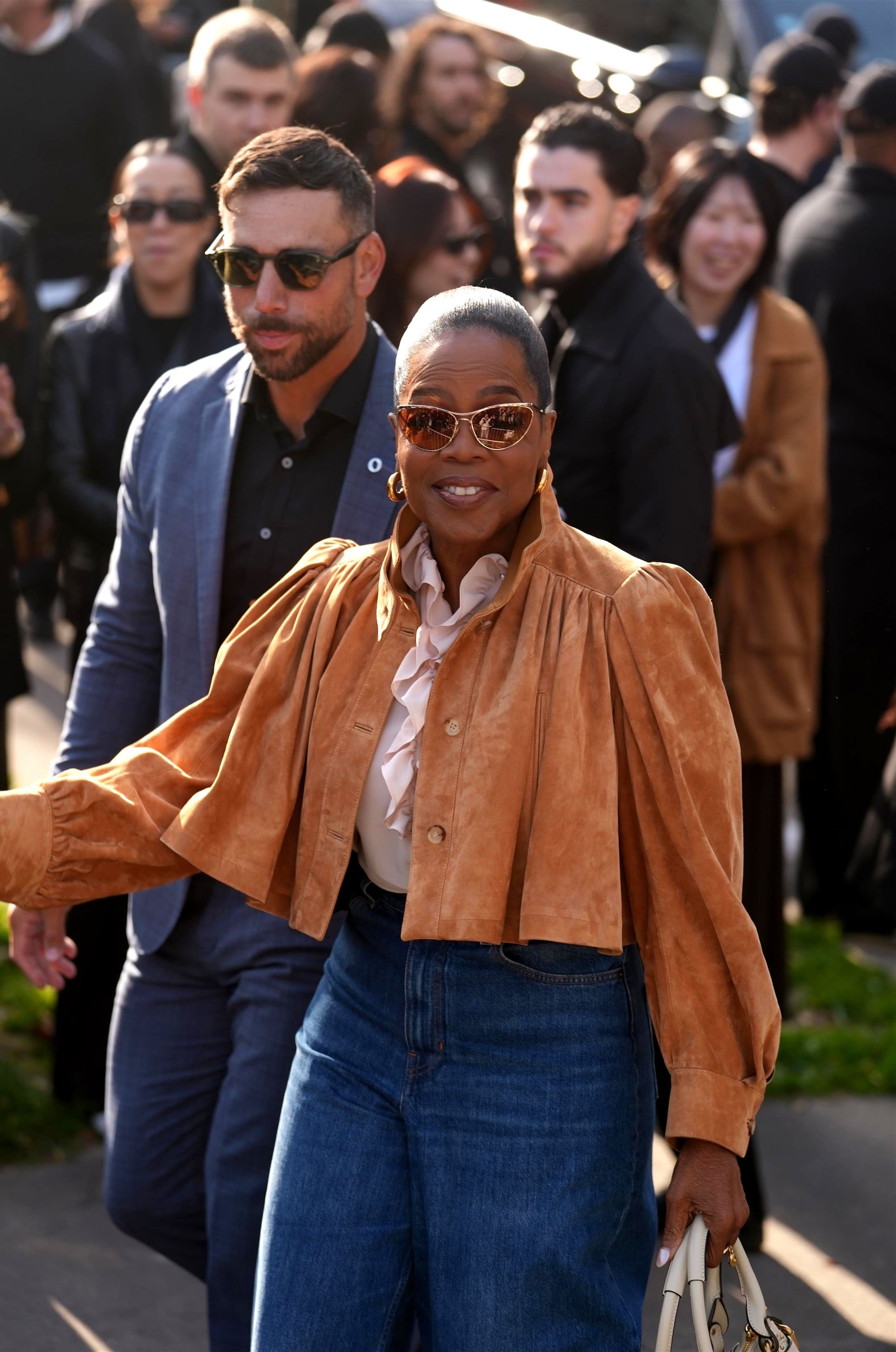 Oprah Winfrey in a tan suede jacket, ruffled white blouse, jeans, and sunglasses at the Chloe Fashion Show.