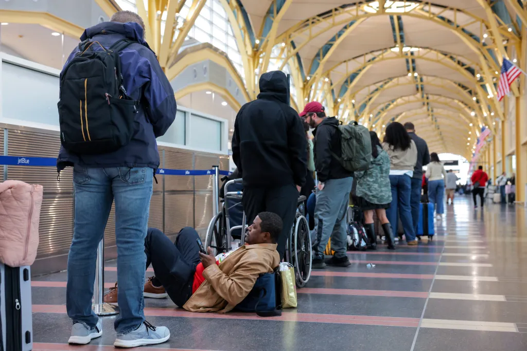 Passengers waiting in a check-in line at Ronald Reagan Washington National Airport, with one man sitting on the floor.