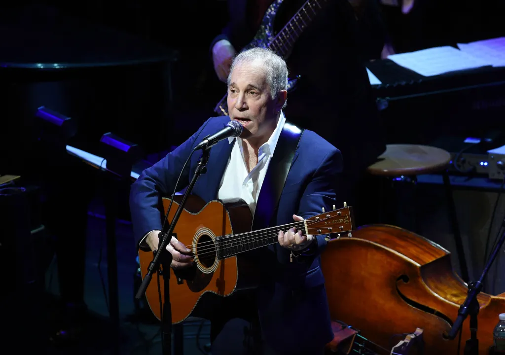 Paul Simon performing on stage, singing into a microphone and playing an acoustic guitar.
