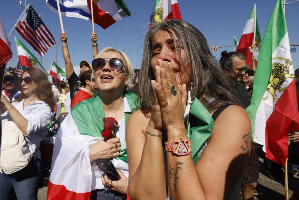 A woman with gray hair covering her mouth with her hands, wearing an Iranian flag and surrounded by other protestors with flags from the US, Israel, and Iran.
