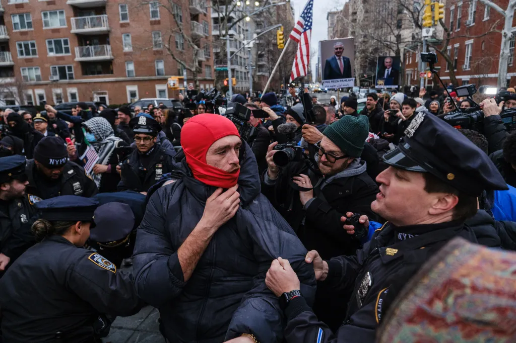 Police officers arresting a protester in a red balaclava, surrounded by a crowd and media.