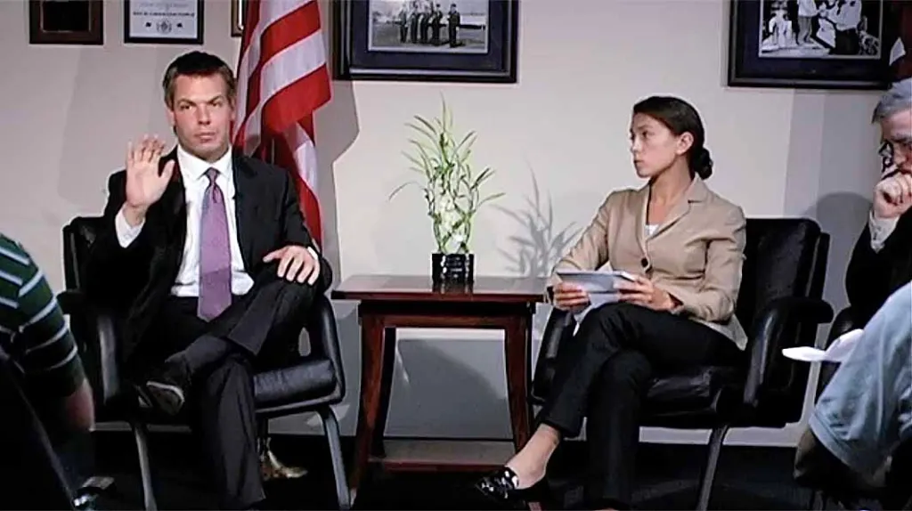 Eric Swalwell seated next to an American flag and a woman holding papers.