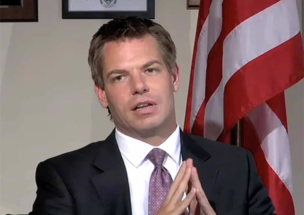 Eric Swalwell, wearing a suit and tie, talks with his hands clasped together, in front of an American flag.