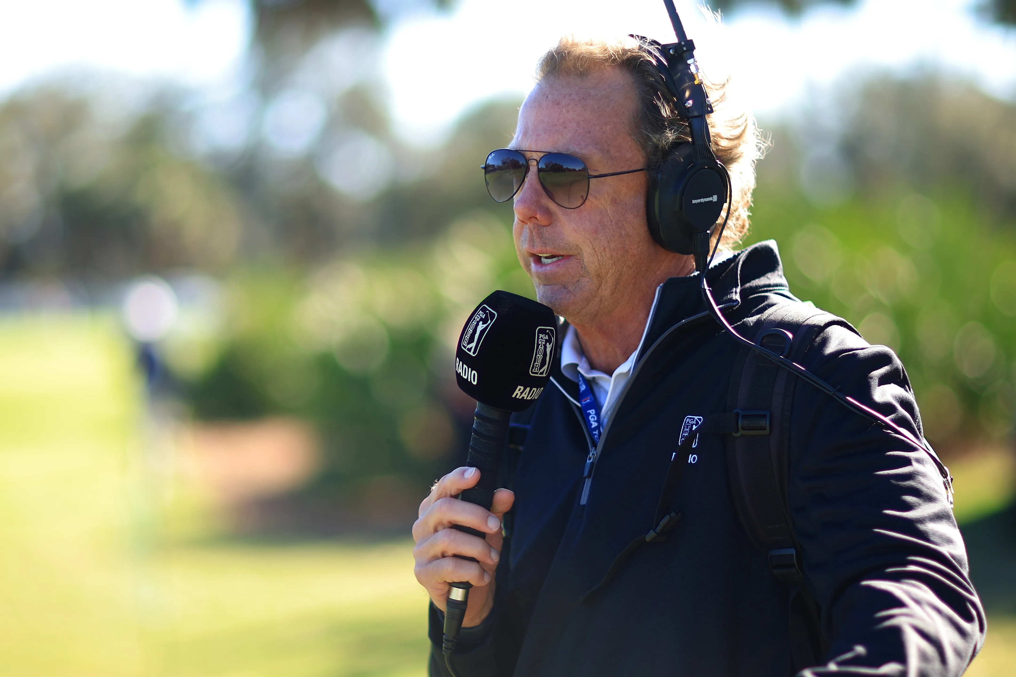 PGA Tour Live announcer Doug Bell wearing sunglasses and headphones, holding a microphone.