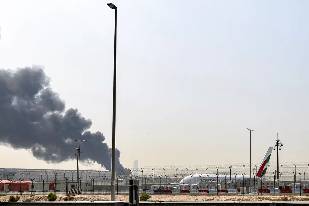 Dark smoke plumes rise from a fire near Dubai International Airport, with a plane and barbed wire fence in the foreground.
