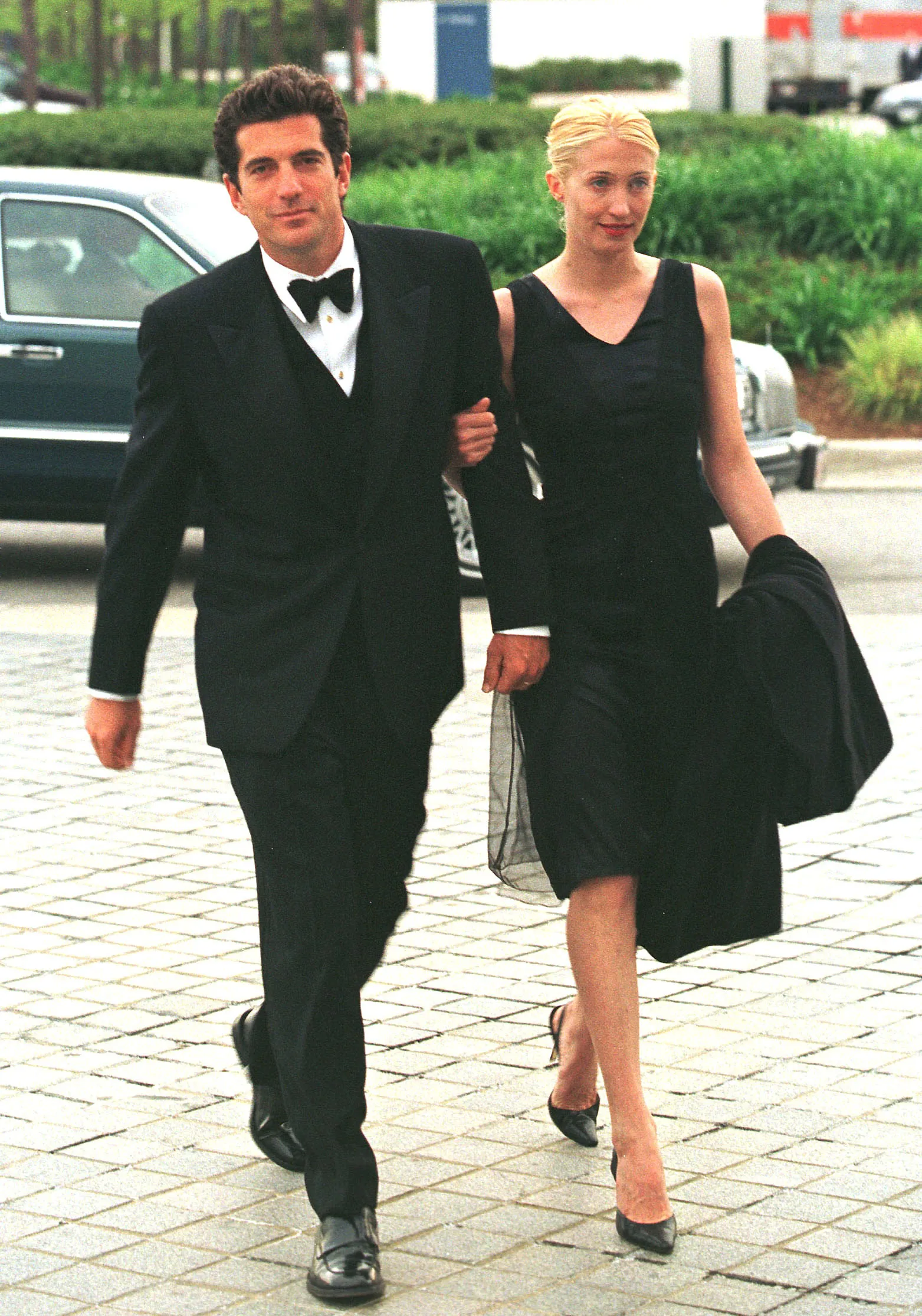 John F. Kennedy Jr. in a tuxedo and Carolyn Bessette-Kennedy in a black dress walk arm-in-arm.