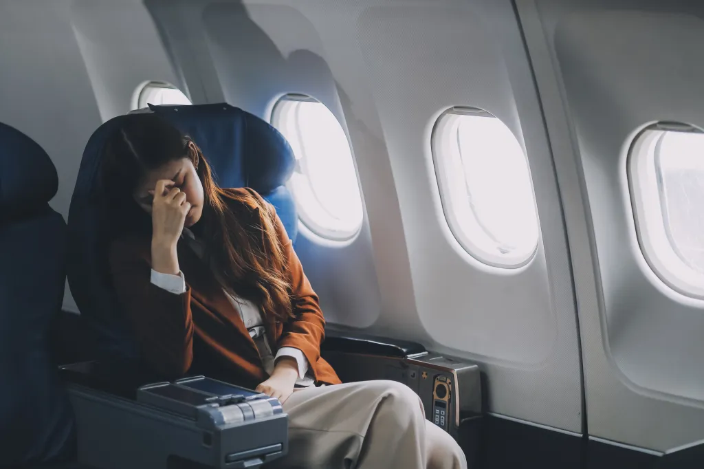 A frustrated woman with long brown hair, wearing a brown jacket, sits in an airplane seat with her head in her hands.