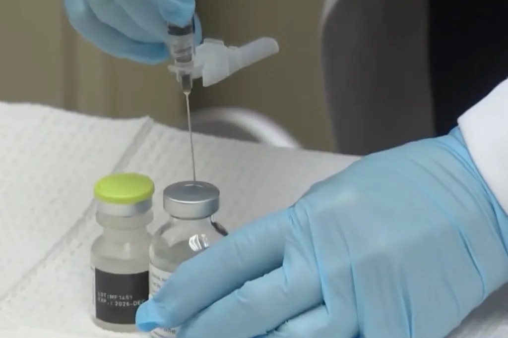 A person in blue gloves draws liquid from a vial with a needle.
