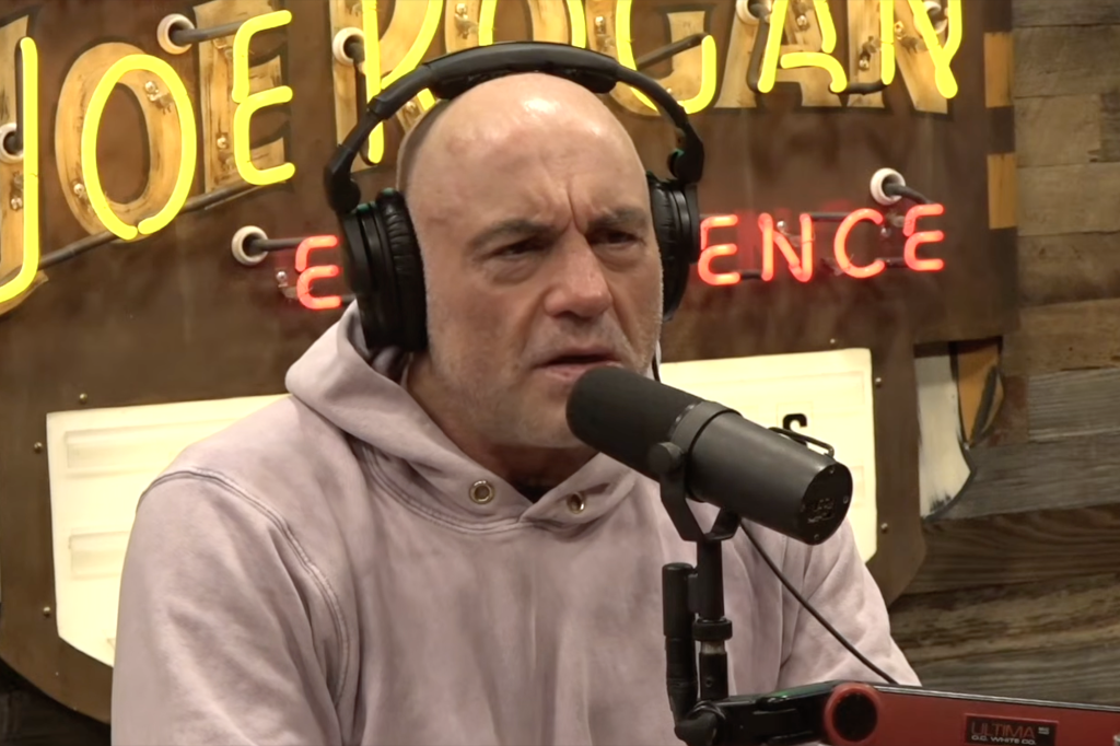 Joe Rogan wearing headphones and speaking into a microphone with neon signs in the background.