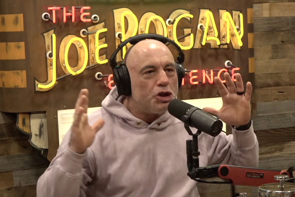 Joe Rogan in a podcast, wearing headphones and a hoodie, gesturing with his hands.
