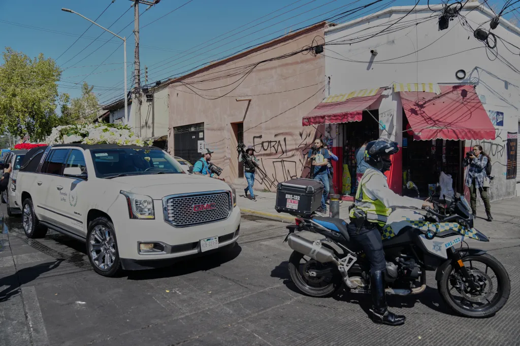A police officer on a motorcycle escorts a white hearse decorated with flowers down a city street.
