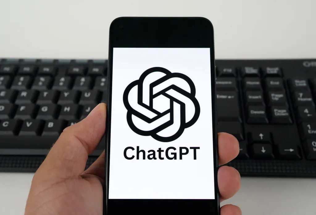 Hand holding a smartphone displaying the ChatGPT logo next to a keyboard.