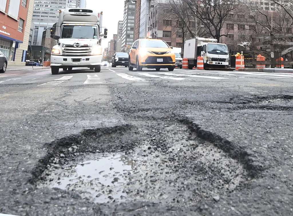 A large pothole with standing water in the foreground of a city street with cars and buildings.