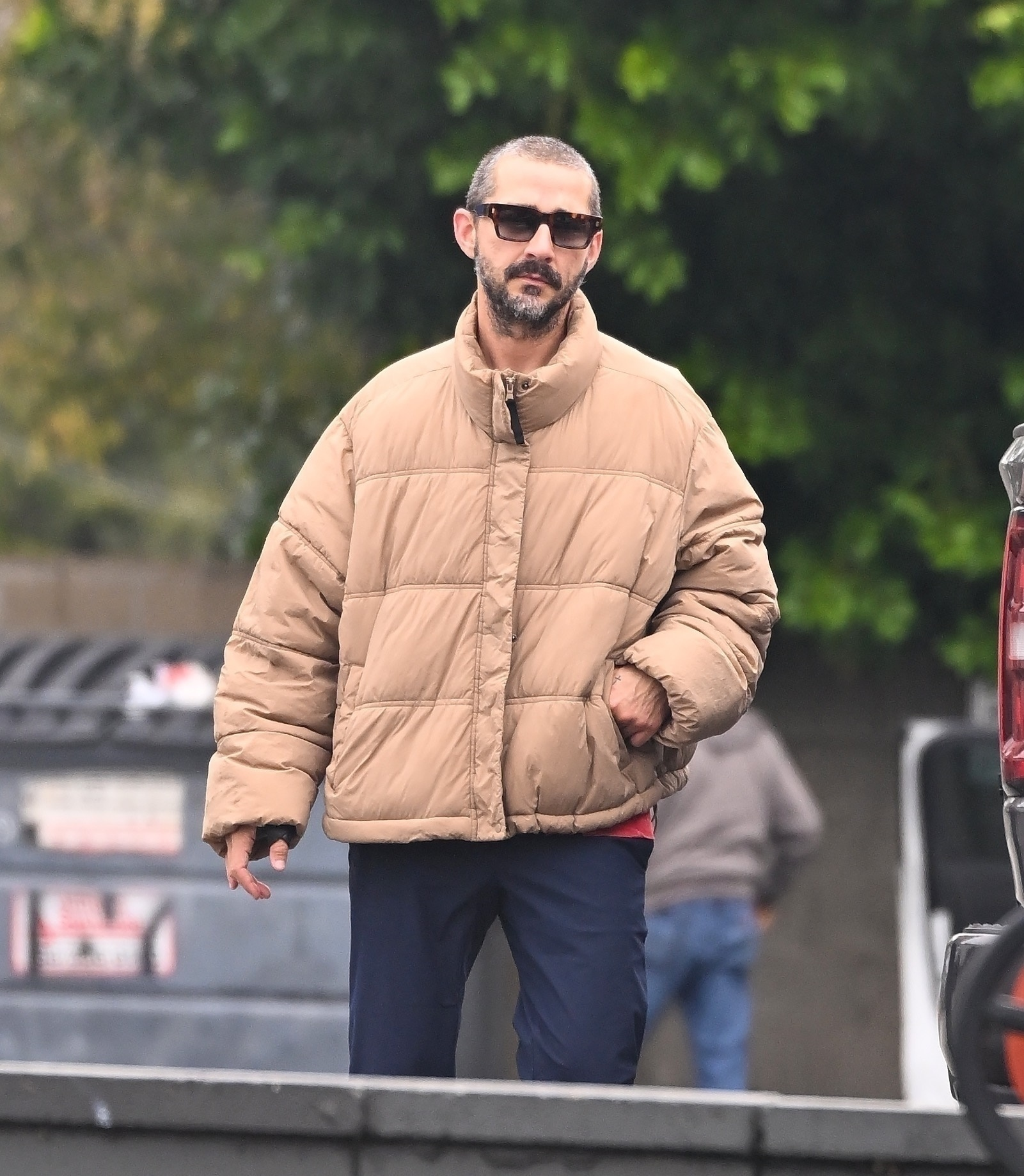Shia LaBeouf walking outside in a tan puffer jacket and sunglasses.