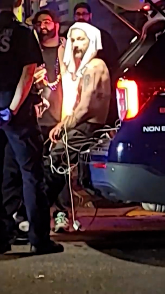 Shia LaBeouf, shirtless and wrapped in a white towel, sits in the back of an ambulance with paramedics nearby.