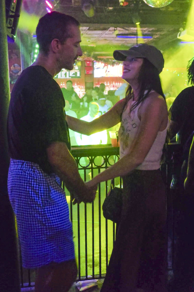 Shia LaBeouf, in a black t-shirt and checked shorts, holding hands with a woman, in a baseball cap, white tank top and black pants, n in a bar.