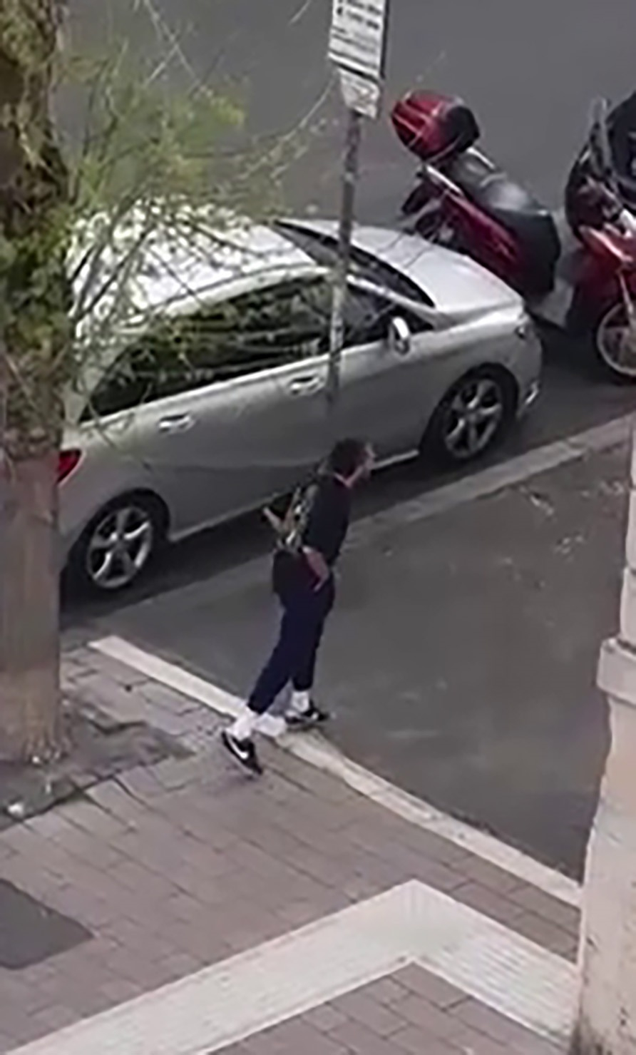Shia LaBeouf walking down a street in Rome, with a silver car and red scooters parked along the curb.