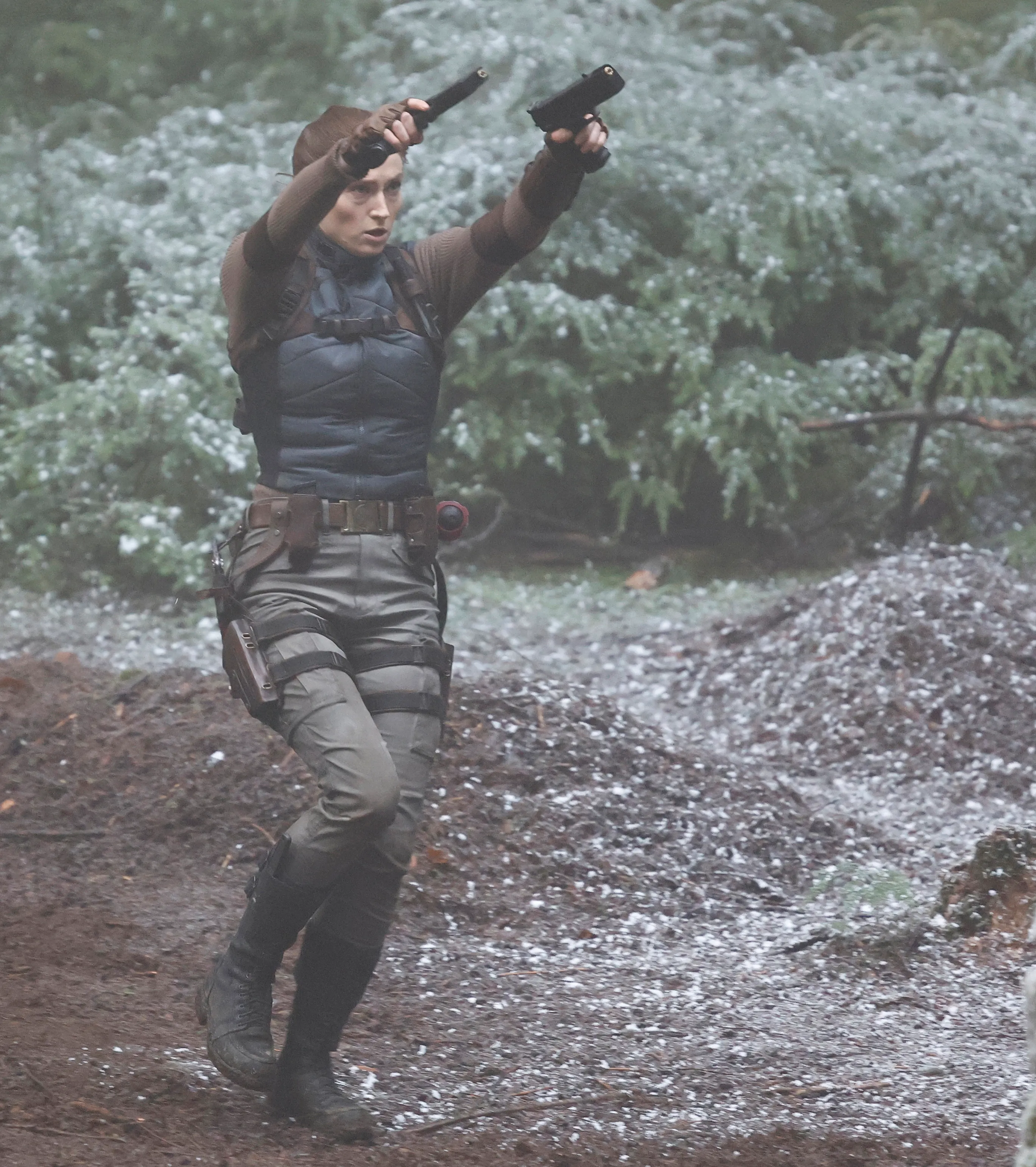 Sophie Turner as Lara Croft holding two guns in a woodland setting.