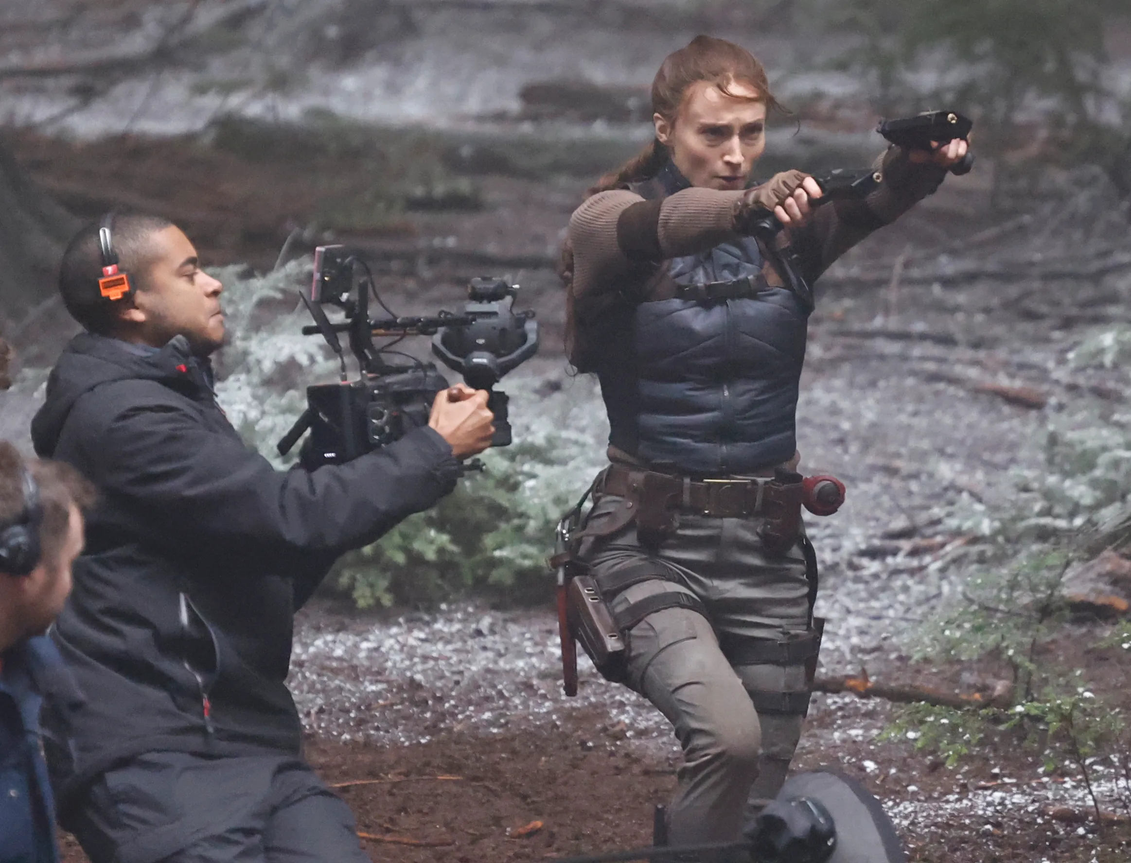 Sophie Turner in character as Lara Croft, aiming two guns in a woodland setting while filming action scenes.