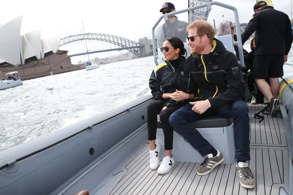 Prince Harry and Meghan Markle sit on a boat on Sydney Harbour, with the Sydney Opera House and Harbour Bridge in the background.