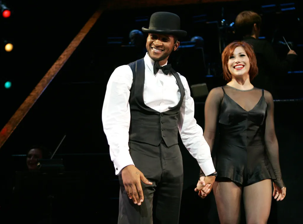 Usher and Bianca Marroquin smiling while holding hands during a curtain call for