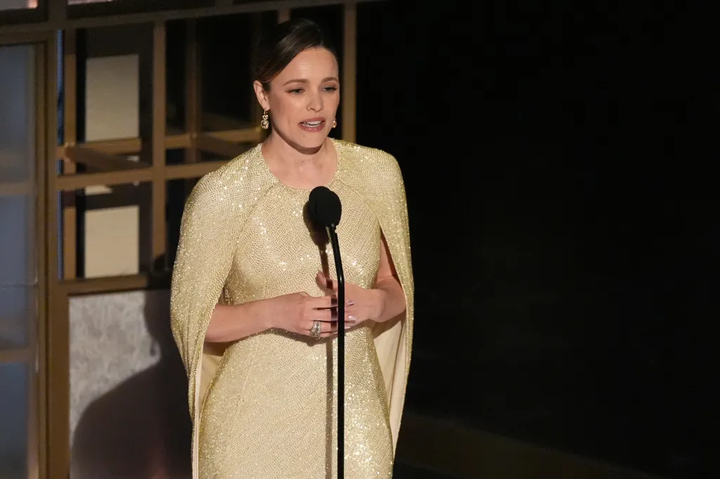 Rachel McAdams speaking at a microphone while wearing a golden gown with a cape-like sleeve design.
