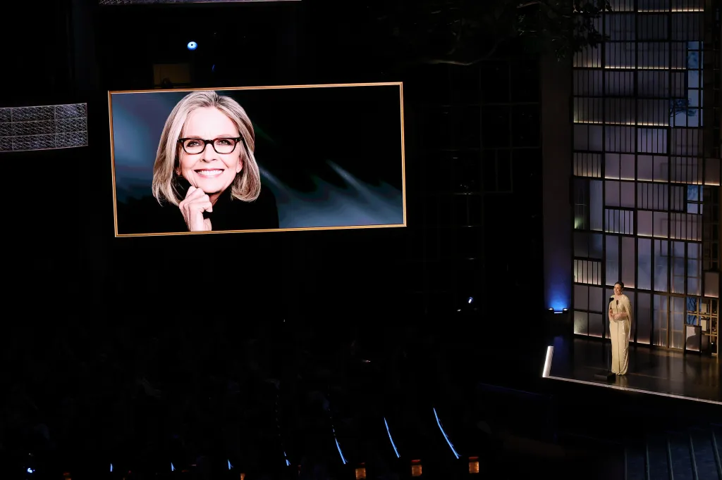 Rachel McAdams speaking onstage during the 98th Oscars, with a large screen displaying a smiling woman in glasses.